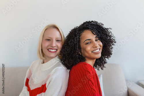 Two beautiful women , blond and african, wearing knitted jumpers sitting on the sofa, back to back and smilling, Cheerful women back against back looking at camera and sitting on the sofa