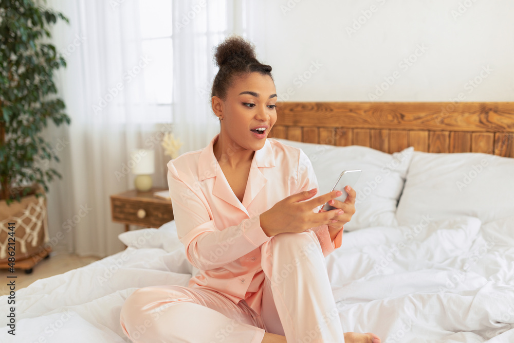 Surprised african american woman readig message on smartphone or browsing internet, sitting on bed at home Surprised african american woman readig message on smartphone or browsing internet, sitting on bed at home