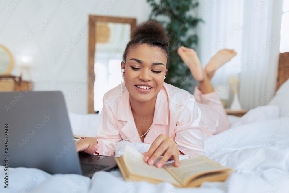 Millennial black lady lying on bed with laptop, reading book and having online class via computer, studying from home Millennial black lady lying on bed with laptop, reading book and having online class via computer, studying from home
