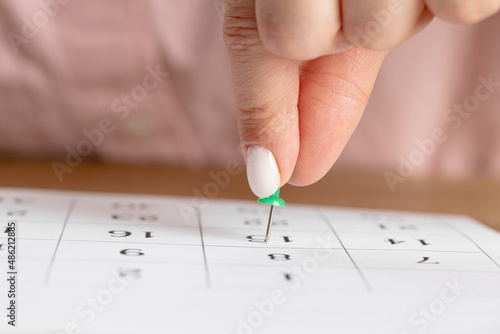 a female hand with a beautiful manicure holds a green pushpin to mark the date on the calendar