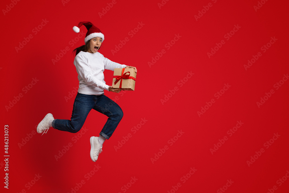 Crazy Sales. Funny Woman Wearing Santa Hat Jumping With Gift In Hands Crazy Sales. Funny Woman Wearing Santa Hat Jumping With Gift In Hands