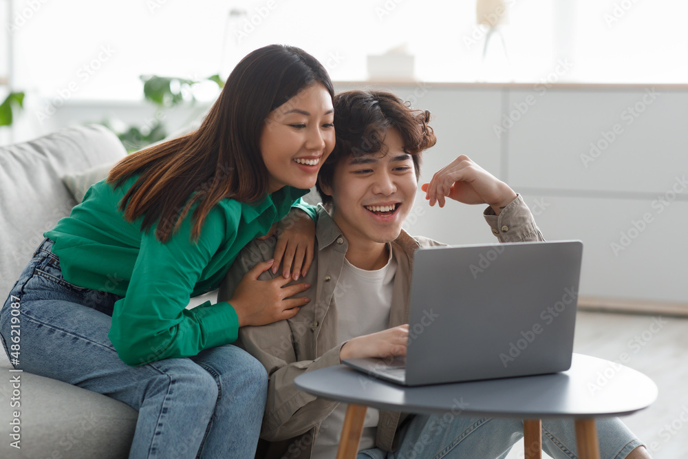 Millennial Asian couple in casual wear using laptop, having online meeting, watching interesting movie in living room Millennial Asian couple in casual wear using laptop, having online meeting, watching interesting movie in living room