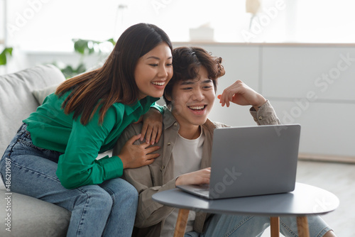 Millennial Asian couple in casual wear using laptop, having online meeting, watching interesting movie in living room