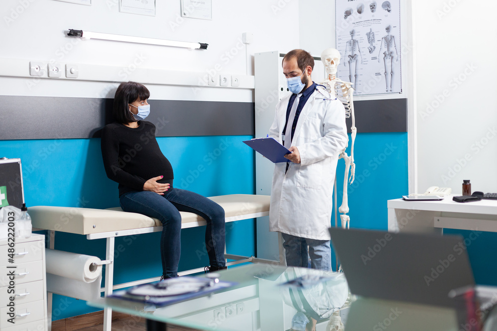 Obraz premium Specialist with face mask consulting pregnant woman in medical cabinet. Patient expecting child and doctor meeting at checkup visit during coronavirus pandemic. Pregnancy examination