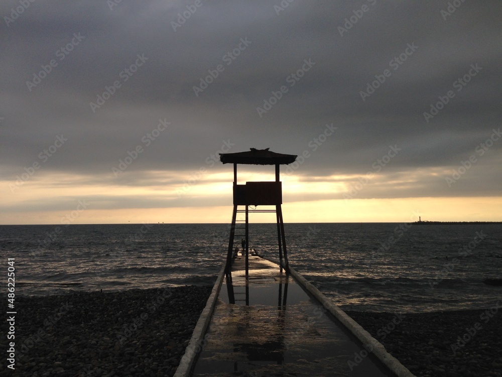 Beach lifeguard post at the sunset.  Lifeguard tower on the stone pier in front of calm sea at the sunset. Rescue. Safety. Water. Measurement. Prevention. Aid. Assistance. Rescuers