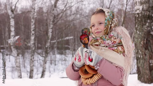 Cute girl in a traditional Russian headscarf and mittens playing on spoons for Maslenitsa festival on winter background. Closeup portrait of a child for Srovetide holiday. photo