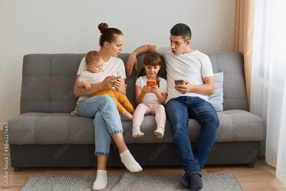 Indoor shot of shocked young family, mother and father looking at daughter's mobile phone with open mouth and surprised facial expression, shock content in kid device. Indoor shot of shocked young family, mother and father looking at daughter's mobile phone with open mouth and surprised facial expression, shock content in kid device.