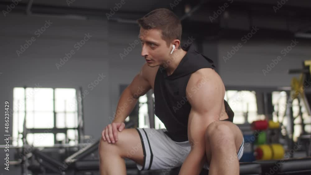 Man with headphones is sitting in gym and lifting dumbbell with one hand, front view. Male athlete trains in gym use dumbbell. Sports concept Man with headphones is sitting in gym and lifting dumbbell with one hand, front view. Male athlete trains in gym use dumbbell. Sports concept