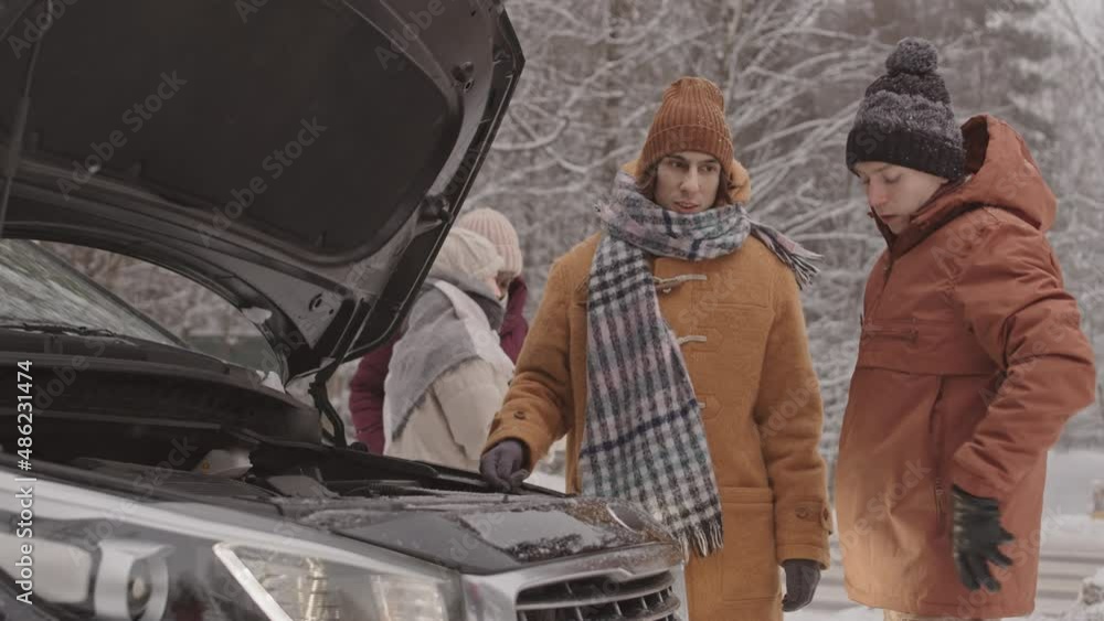 Medium slowmo shot of two young Caucasian men looking at opened car bonnet trying to find out problem standing outdoors in forest on snowy winter day while their girlfriends chatting in background Medium slowmo shot of two young Caucasian men looking at opened car bonnet trying to find out problem standing outdoors in forest on snowy winter day while their girlfriends chatting in background