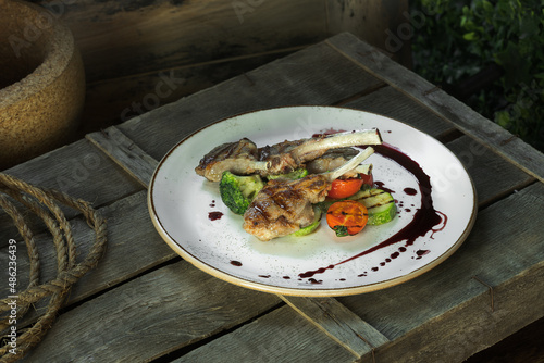 Fototapeta Naklejka Na Ścianę i Meble -  grilled rack of lamb and garnished with drops of sauce lies on a white dish along with tomatoes, zucchini and broccoli. The dish stands on a wooden box, rustic style