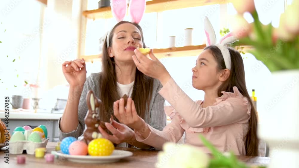 Close up happy mother, daughter in the kitchen during breakfast, eating delicious chocolate rabbit and colorful Easter eggs. Happy Easter atmosphere. Celebrating spring holiday concept. Close up happy mother, daughter in the kitchen during breakfast, eating delicious chocolate rabbit and colorful Easter eggs. Happy Easter atmosphere. Celebrating spring holiday concept.