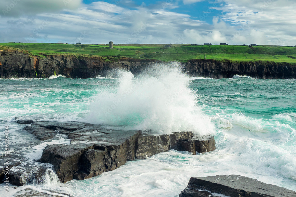 Powerful ocean wave crushes on rock, cliff with green fields and blue cloudy sky in the background. Beautiful nature scenery. Doolin, county Clare, Ireland. Irish landscape.