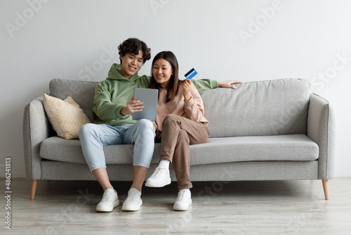 Young Asian couple with credit card and tablet sitting on sofa, ordering goods on internet, buying things in web store