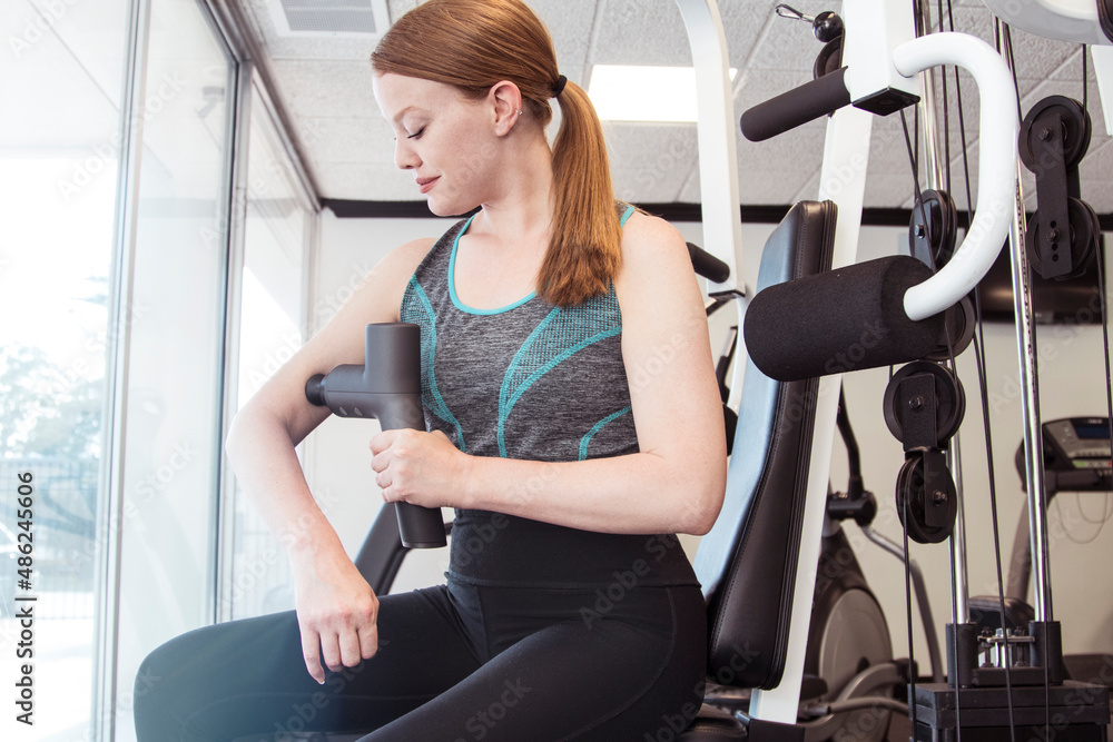Fototapeta premium Young beautiful redhead woman using massage gun on arm in gym