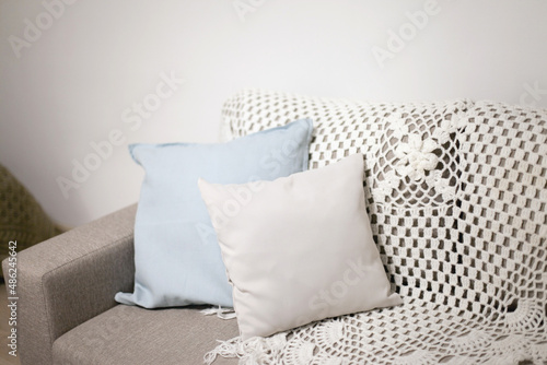 Fototapeta Naklejka Na Ścianę i Meble -  Beige couch at home with gray and blue pillow and crochet blanket, cozy sofa in apartment home. Couch decor in neutral colors interior at home.