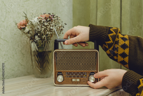 In a light green interior, the hands of a man in a knitted sweater with a pattern put a radio receiver on a chest of drawers and switches a musical composition, a bouquet of flowers stands behind.