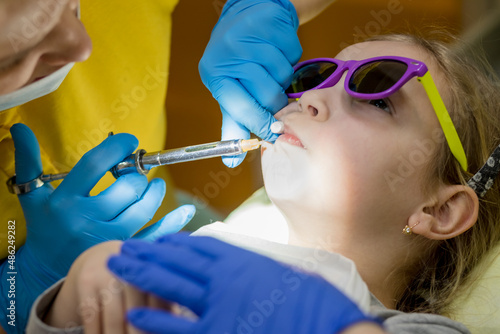 side view of dentist holding a syringe and anesthetizing her little girl patient. Doctors dentists giving for child an injection anesthesia. Stomatology for kids concept.comfortable dental treatment
