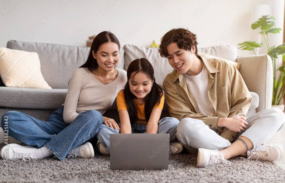 Smiling young asian female, male and teen girl sitting on floor and watching video, searching, browsing on laptop Smiling young asian female, male and teen girl sitting on floor and watching video, searching, browsing on laptop