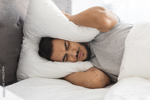 Insomnia Concept. Annoyed Middle Eastern Guy Covering Ears With Pillow photo