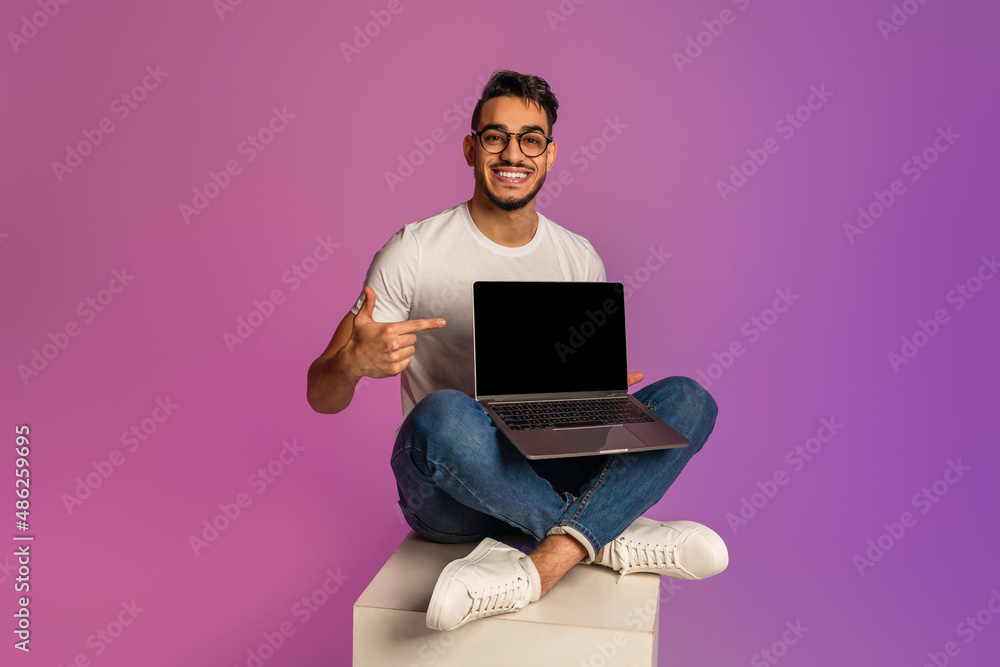Happy Arab man sitting cross legged with laptop, pointing at blank screen, showing space for mockup in neon light Happy Arab man sitting cross legged with laptop, pointing at blank screen, showing space for mockup in neon light