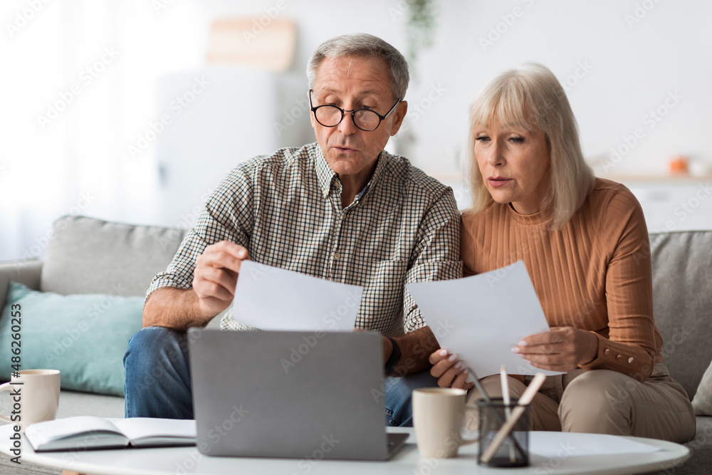 Senior Spouses Reading Papers And Bills Sitting Near Laptop Indoors Senior Spouses Reading Papers And Bills Sitting Near Laptop Indoors