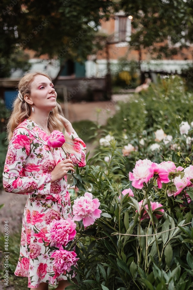 Beautiful cheerful young blonde girl wearing summer dress standing isolated over pink background, holding bouquet of peonies Beautiful cheerful young blonde girl wearing summer dress standing isolated over pink background, holding bouquet of peonies