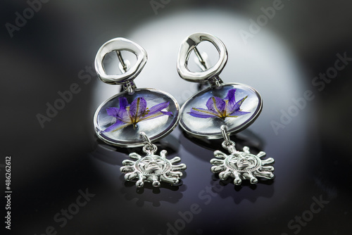 silver earrings with a living flower in glass and a pendant in the form of the sun lie on a mirror surface, close-up, dark background