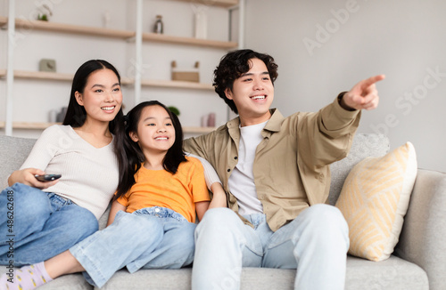 Smiling japanese young man shows on empty space with hand to wife and teen girl, sit on sofa in living room © Prostock-studio