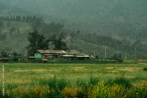 Village house against the backdrop of forest and mountains, field in front of the house, log house