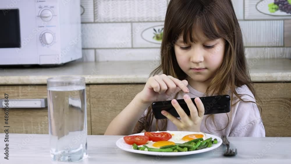 The child does not want to eat breakfast.The girl in the kitchen refuses to eat and plays on the phone.Internet addiction.A child and a gadget. Social problems.Fried eggs, tomatoes and beans on a plat The child does not want to eat breakfast.The girl in the kitchen refuses to eat and plays on the phone.Internet addiction.A child and a gadget. Social problems.Fried eggs, tomatoes and beans on a plat