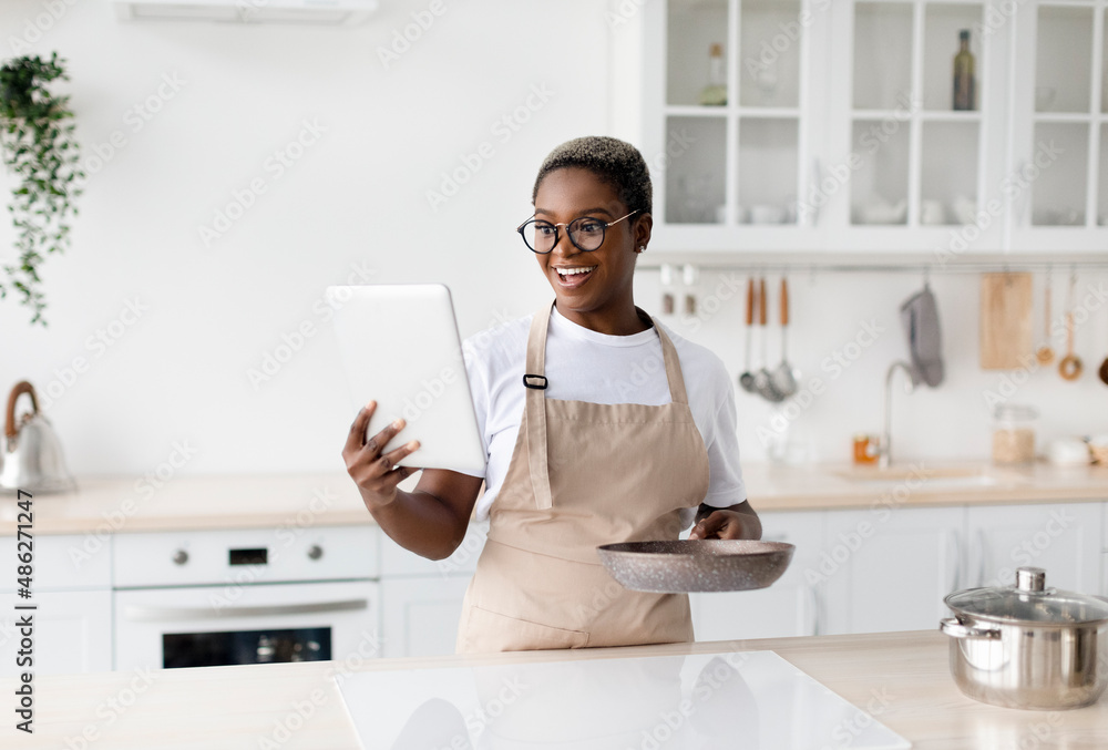 Surprised happy young african american woman in apron and glasses looks at tablet and prepares dinner