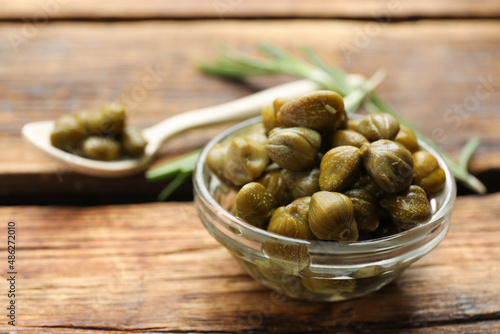 Tasty capers in glass bowl on wooden table, closeup. Space for text
