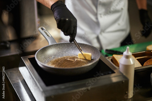 Crope view of chef's hands in black gloves melting piece of butter in frying pan. Male in chef uniform using tweezers for cooking with ingredients and equipment on background. Concept of cooking. 