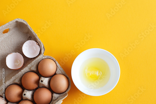 raw eggs in a package next to white bowl with one broken egg in it on a yellow background