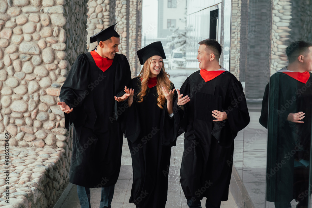 Graduate friends in gowns walk along university corridor and talking excitedly