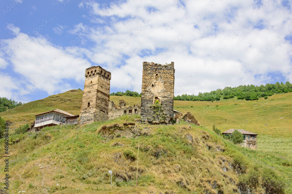 Settlement with two medieval svan towers on the road to Ushguli medieval fortified village in Svanetia, Georgia. Green mountain slopes, ols yellow and grey stone walls, blue sky with clouds