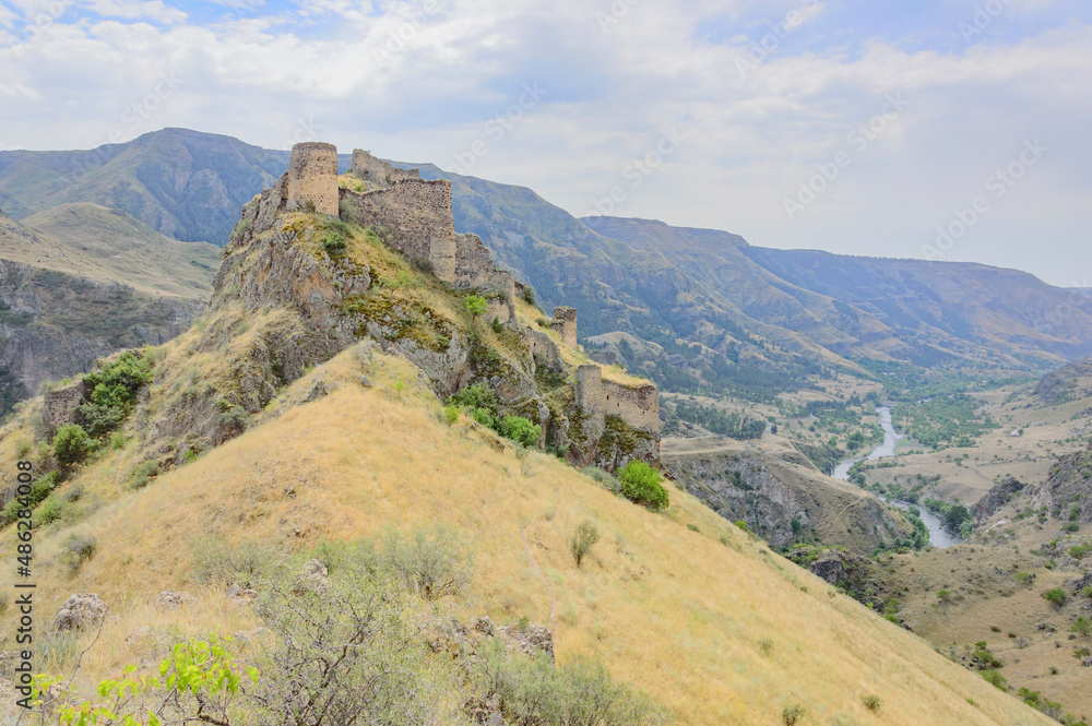 Ruined medieval Tmogvi fortress and Caucasian mountains in southern Georgia. Ancient stone walls and towers, dry yellow grass, green trees, rocky slopes, blue sky