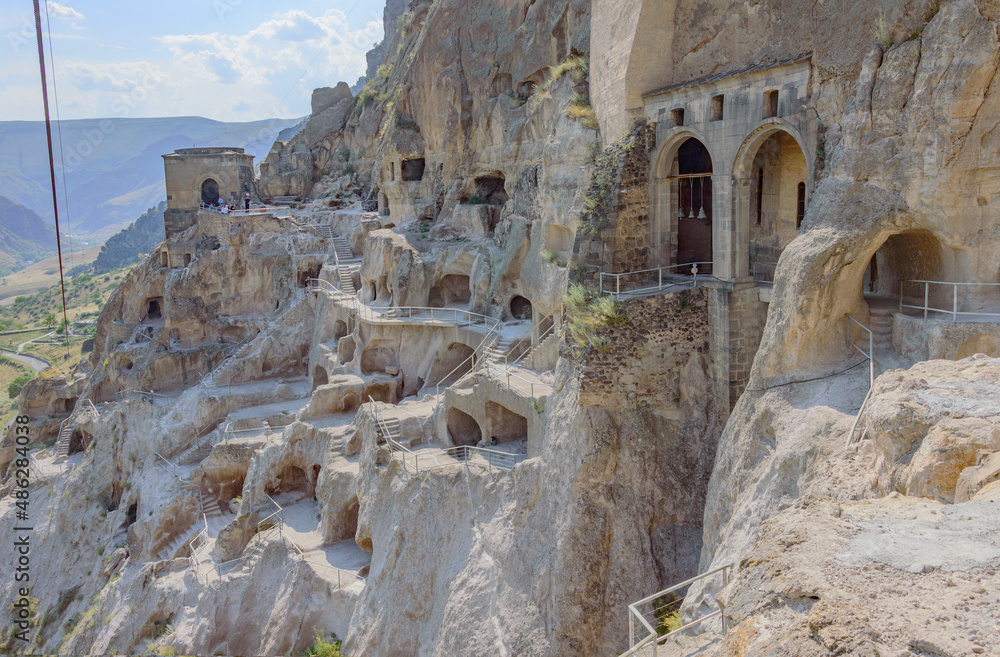 Vardzia ruined cave city in Georgia. Grey and yellow rock cliff with numerous caves, green nountain slopes, blue sky with clouds