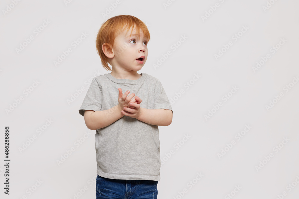 Beautiful portrait of a red-headed boy and child in grey T-shirt on grey or white background looking surprised to the right side. Standing isolated looking adorable. Copy space. Beautiful portrait of a red-headed boy and child in grey T-shirt on grey or white background looking surprised to the right side. Standing isolated looking adorable. Copy space.