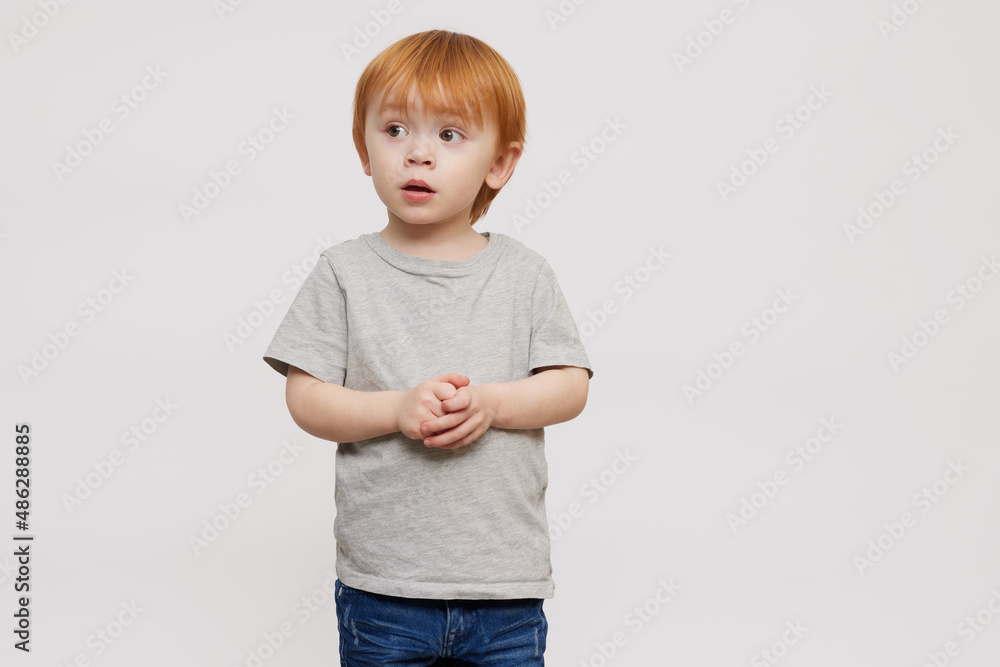 Cute red-haired and red-headed boy in grey T-shirt on grey or white background folding his hands and looking a bit scared to the left side. Standing isolated looking adorable. Copy space. Cute red-haired and red-headed boy in grey T-shirt on grey or white background folding his hands and looking a bit scared to the left side. Standing isolated looking adorable. Copy space.
