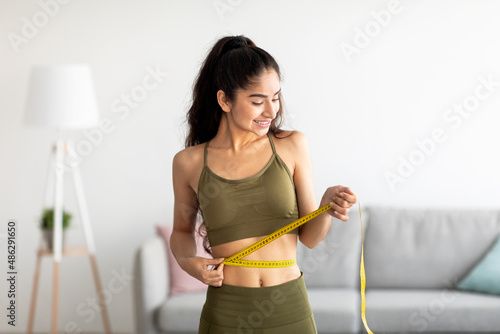 Successful weight loss concept. Fit young Indian woman in sports clothes measuring waist with tape at home photo
