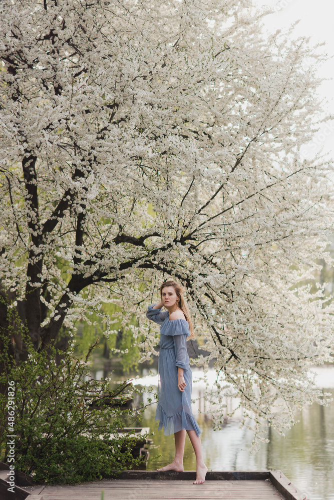 Young pretty caucasian blonde woman with freckles wearing natural makeup in light blue dress near the beautiful blooming spring tree by the lake. Youth, freshness, beauty, happiness, emotions concept.