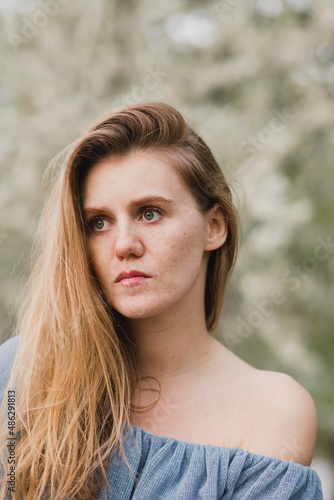 Young pretty caucasian blonde woman with freckles wearing natural makeup in light blue dress near the beautiful blooming spring tree by the lake. Youth, freshness, beauty, happiness, emotions concept.