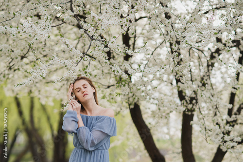 Young pretty caucasian blonde woman with freckles wearing natural makeup in light blue dress near the beautiful blooming spring tree by the lake. Youth, freshness, beauty, happiness, emotions concept.