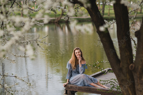 Young pretty caucasian blonde woman with freckles wearing natural makeup in light blue dress near the beautiful blooming spring tree by the lake. Youth, freshness, beauty, happiness, emotions concept. © anna_gorbenko