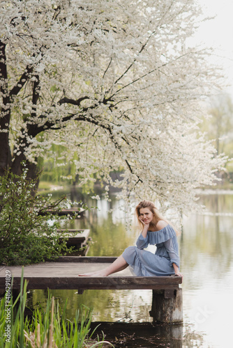 Young pretty caucasian blonde woman with freckles wearing natural makeup in light blue dress near the beautiful blooming spring tree by the lake. Youth  freshness  beauty  happiness  emotions concept.