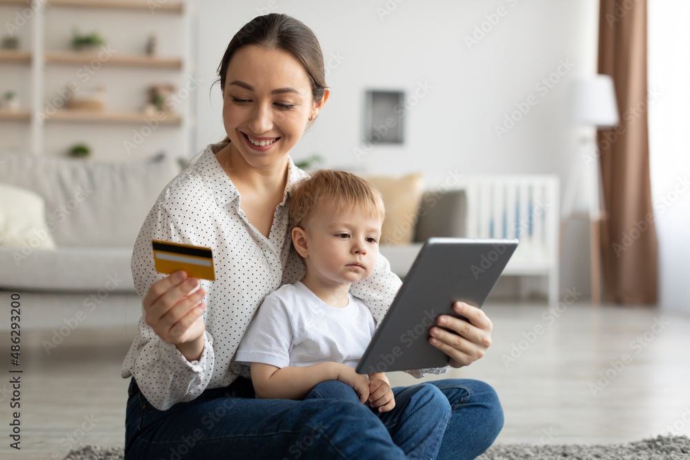 Young mother with child boy using digital tablet and credit card for internet purchases, sitting on floor in living room Young mother with child boy using digital tablet and credit card for internet purchases, sitting on floor in living room