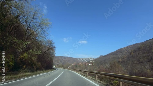 Slow driving of a car or motorcycle on a winding mountain road surrounded by trees. A mountain village in the distance. Autumn, blue clear sky. POV shot photo