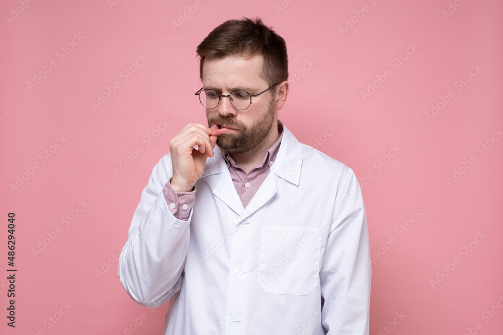 Doctor in a white coat pulls his lip with his fingers and looks upset. Doctor in a white coat pulls his lip with his fingers and looks upset.