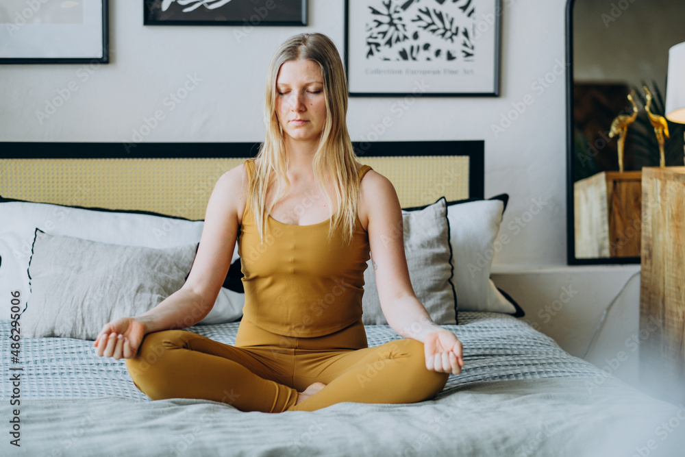 Beautiful blonde woman meditates while sitting in the lotus position, concentration and affirmation in bed in the bedroom. Stream state Beautiful blonde woman meditates while sitting in the lotus position, concentration and affirmation in bed in the bedroom. Stream state